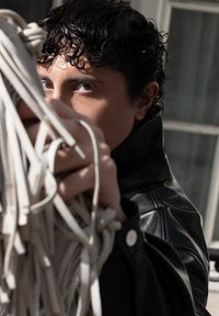 Zwarte leren jas met een hoge kraag; persoon houdt lichtgekleurde, gestructureerde touwen of koorden vast; zonlicht benadrukt de details.