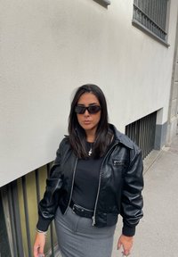 Μαύρη δερμάτινη καμπαρντίνα με φερμουάρ μπροστά, ραμμένα μανίκια και μια μικρή τσέπη. Φορεμένη πάνω από μια εφαρμοστή μαύρη μπλούζα και γκρι μολυβένια φούστα. Μαύρα γυαλιά ηλίου.