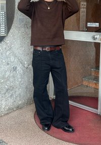 Suéter marrón con textura, vaqueros negros de pierna ancha, cinturón marrón y zapatos negros, de pie junto a una pared de piedra y una puerta de cristal.