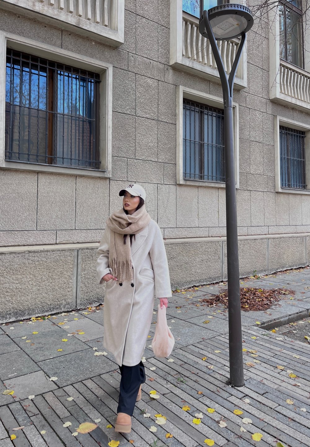 Beige Wolle Mantel mit schwarzen Knöpfen, hellbeige Schal mit Fransen, schwarze Hose und hellbeige Plateaustiefel. Geht entlang eines mit Blättern bedeckten Gehwegs.