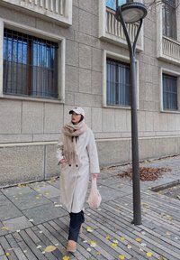 Beige Wolle Mantel mit schwarzen Knöpfen, hellbeige Schal mit Fransen, schwarze Hose und hellbeige Plateaustiefel. Geht entlang eines mit Blättern bedeckten Gehwegs.