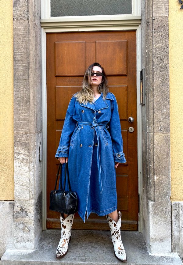 Femme portant un long manteau en jean bleu, des lunettes de soleil, tenant un sac à main noir et debout devant une porte en bois marron avec un encadrement en pierre.