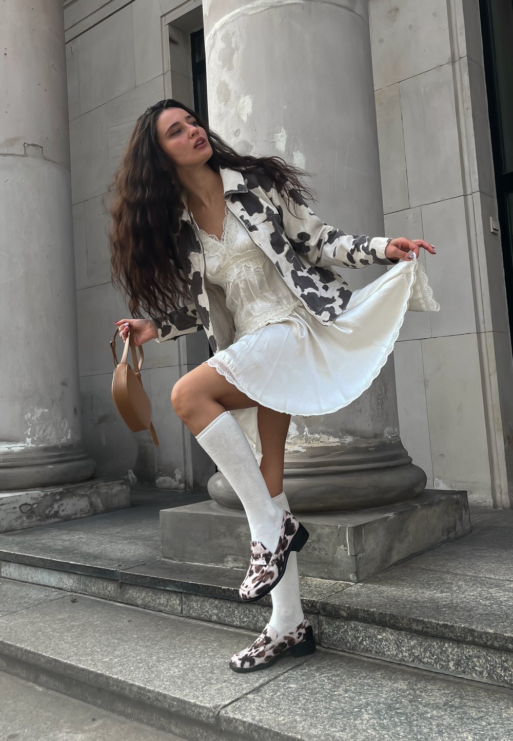 Model wearing a cow-print jacket over a white lace dress, with knee-high white socks and patterned shoes, standing on stone steps beside columns.