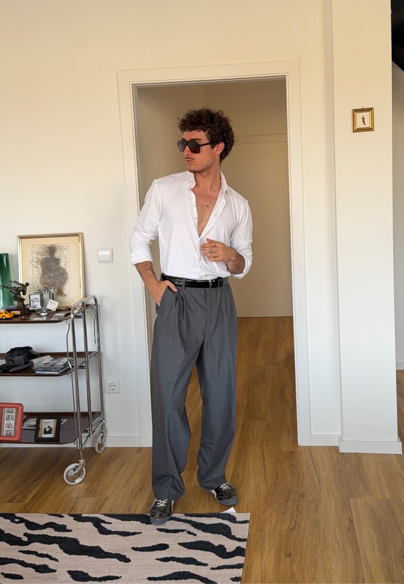 Mann mit lockigem Haar, der eine Sonnenbrille, ein weisses Hemd und graue Hosen trägt, steht mit einer Hand in der Tasche in einem Raum mit Holzboden.