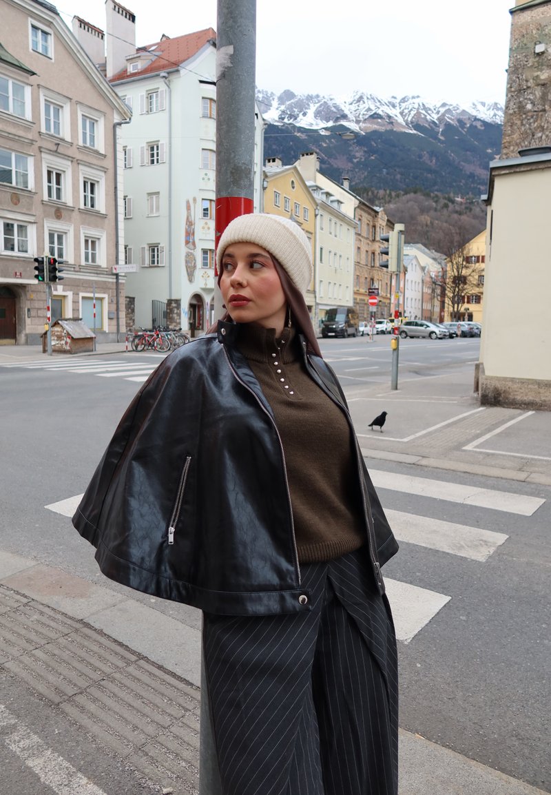 Mujer con un gorro beige, chaqueta de cuero negra y pantalones a rayas, de pie junto a un poste en una calle de la ciudad con montañas cubiertas de nieve al fondo.