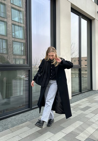 Abrigo negro largo sobre una camisa negra abotonada, jeans grises y zapatos gruesos negros. Textura suave, colores neutros y líneas limpias. Entorno urbano moderno.