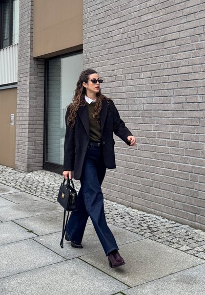 Blazer negro de gran tamaño, suéter verde oliva sobre camisa blanca, vaqueros oscuros de pierna ancha, bolso negro, botas de tacón oscuro, sobre una acera de piedra.
