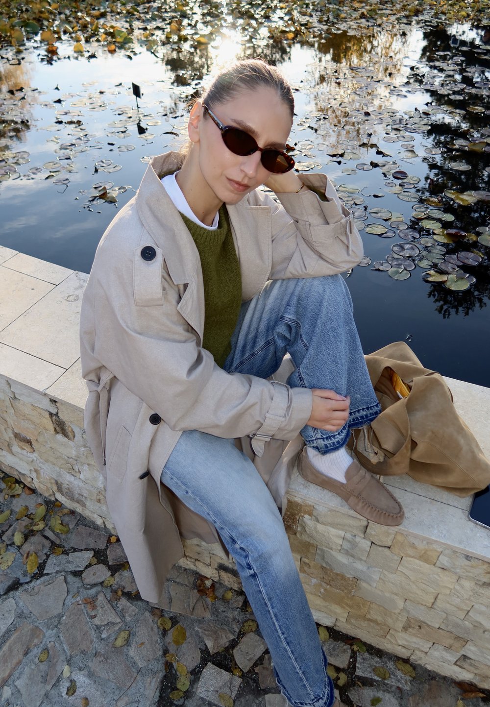 Béžový trenčkot s veľkými gombíkmi, zelený sveter, svetlomodré džínsy a hnedé nazúvacie topánky. Sedí pri vode s lekno listami.