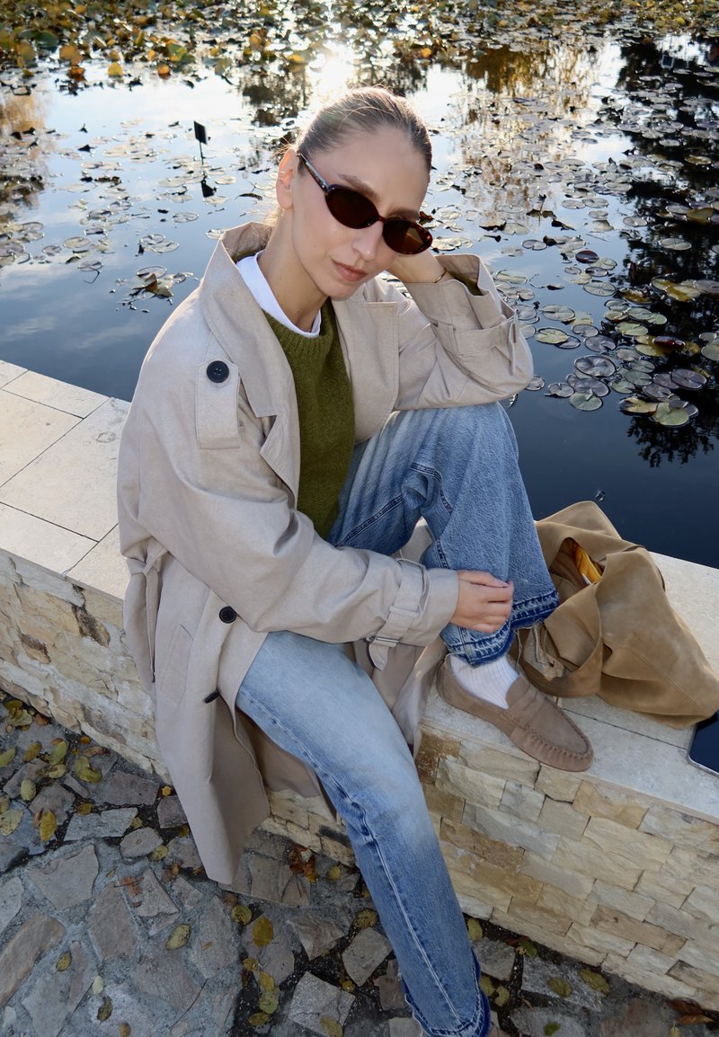 Casaco trench bege com botões grandes, suéter verde, jeans azul claro e sapatos deslizantes castanhos. Sentado junto à água com nenúfares.