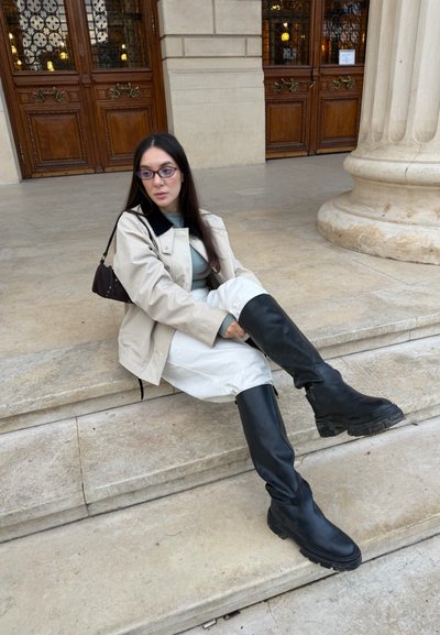Mujer con el pelo largo y oscuro y gafas sentada en unos escalones de piedra, vistiendo una chaqueta beige, pantalones claros, botas negras hasta la rodilla y llevando un pequeño bolso negro.