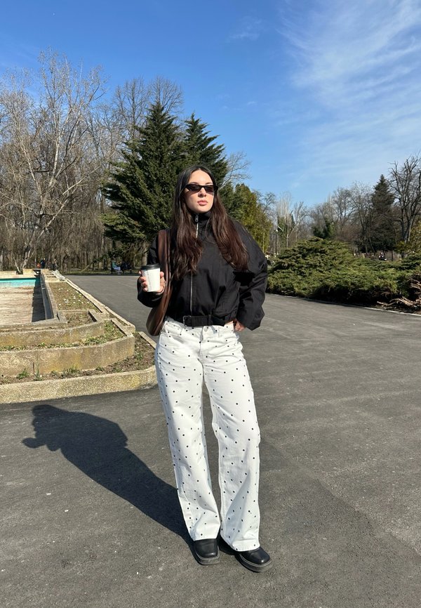 Femme portant une veste noire, un pantalon blanc à pois et des lunettes de soleil, tenant un gobelet à emporter, debout sur un chemin pavé dans un parc.