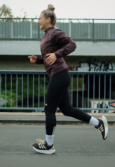 Ropa de correr que incluye una chaqueta ligera morada, leggings negros con bolsillos laterales y zapatillas deportivas marrones con suelas y cordones blancos.