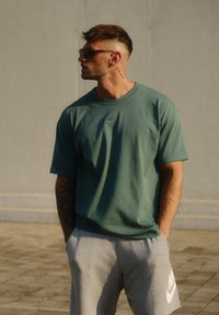 Zöld pamut póló kicsi Nike logóval, szürke melegítőnadrággal, amelyen fehér Nike logó látható. Hétköznapi öltözet minimális részletezéssel.