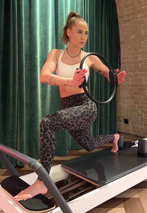 Žena u leopard uzorku helanki i bijelom sportskom grudnjaku balansira na Pilates reformer stroju držeći Pilates prsten s fokusiranim izrazom lica.