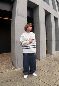 Λευκή μπλούζα με λαιμόκοψη και εκτυπωμένο κείμενο με ναυτικές λεπτομέρειες, συνδυασμένη με χαλαρά μπλε τζιν και λευκά αθλητικά παπούτσια, μπροστά σε γκρίζα δομή.