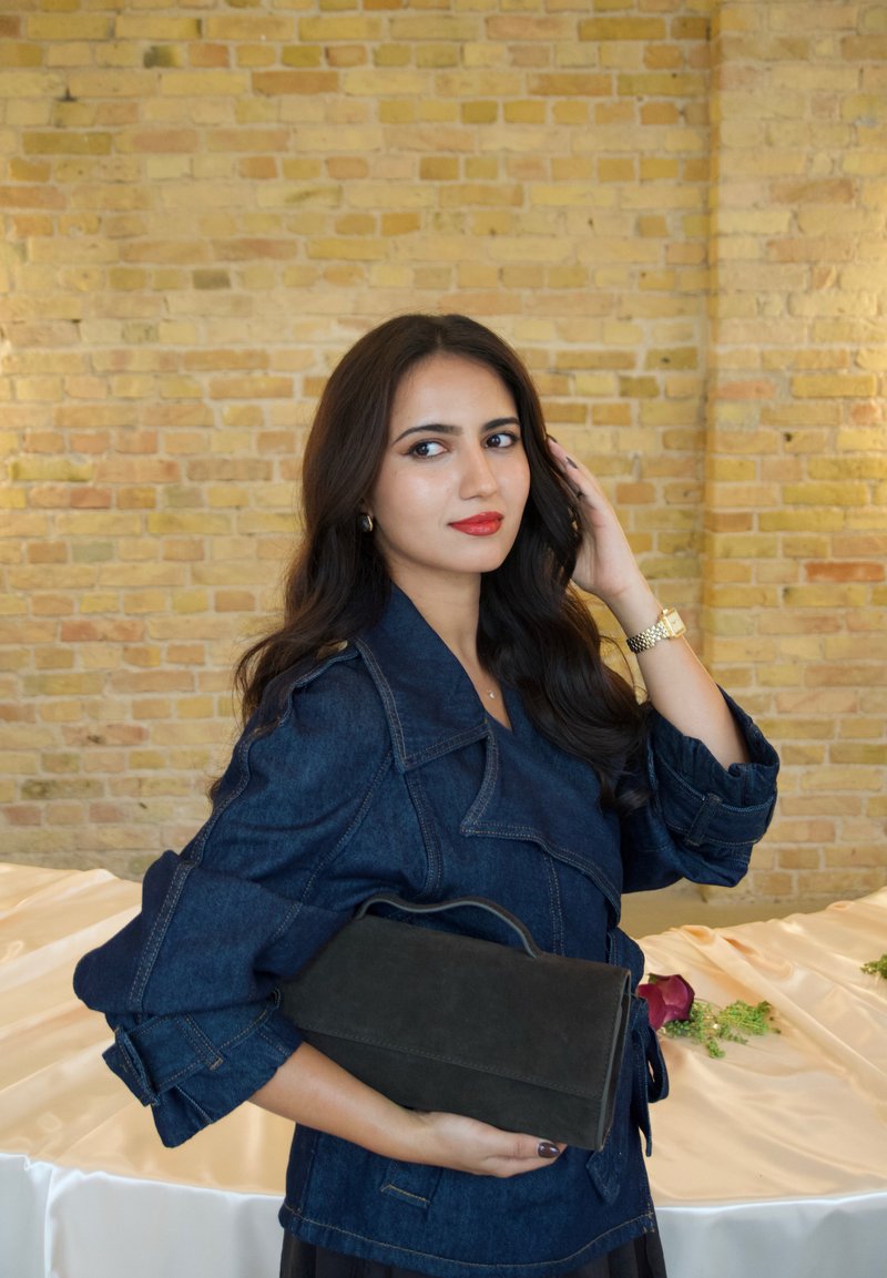 Sac de soirée en daim noir, de forme rectangulaire structurée et avec une poignée supérieure, tenu par une personne portant une veste en jean, sur un fond clair.