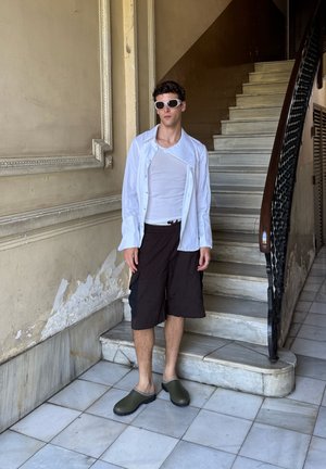 Camisa blanca de botones con rayas, camiseta blanca sin mangas, pantalones cortos negros y zuecos verdes; de pie en escaleras blancas de azulejos con barandilla ornamentada.