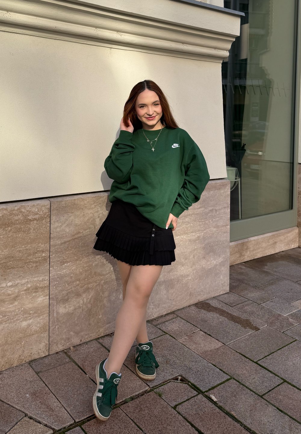 Jovem mulher com um sweatshirt verde da Nike, saia preta em camadas e ténis verdes, encostada a uma parede de pedra ao ar livre, sorrindo.