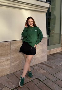 Jovem mulher com um sweatshirt verde da Nike, saia preta em camadas e ténis verdes, encostada a uma parede de pedra ao ar livre, sorrindo.