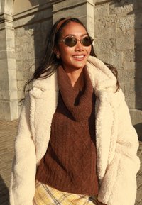 Kreemjas fliisist jakk pruuni soonikkoega kaelusega kudumi kohal, koos ruudulise seeliku ja ümarate päikeseprillidega. Tekstuuriga, soojad materjalid.