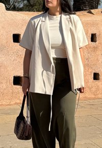 Femme portant un blazer blanc à manches courtes par-dessus un haut blanc et un pantalon vert olive, tenant un sac à main tissé foncé, debout sur un sol pavé.