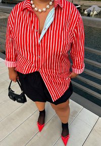 Red and white striped button-up shirt worn over a black skirt, complemented by black socks and vibrant red pointed shoes. Accompanied by a small black handbag.
