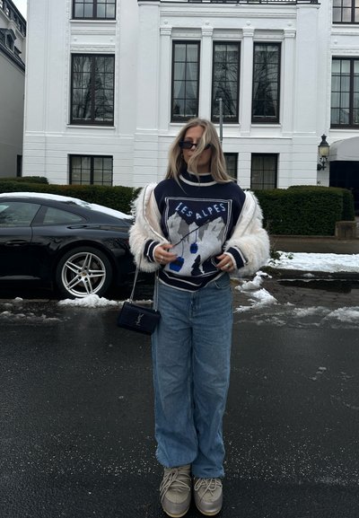 Suéter navy con diseño "LES ALPES", chaqueta de piel sintética clara, jeans azul y zapatos beige. Bolso negro con cadena plateada. Nieve visible en el suelo.