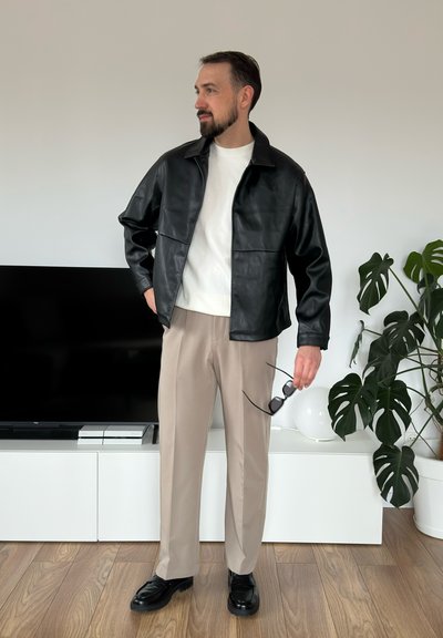 Chaqueta de cuero negra, suéter blanco, pantalones beige y zapatos negros. Sosteniendo gafas de sol, de pie frente a una pared neutral y plantas.