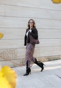 Cardigã castanho, suéter branco, saia xadrez, botas de salto alto pretas, carregando uma bolsa castanha, a caminhar por um caminho de pedras contra uma parede clara.