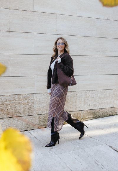 Cardigan marrón, suéter blanco, falda de cuadros, botas de tacón negro, llevando un bolso marrón, caminando por un camino de piedra contra una pared clara.