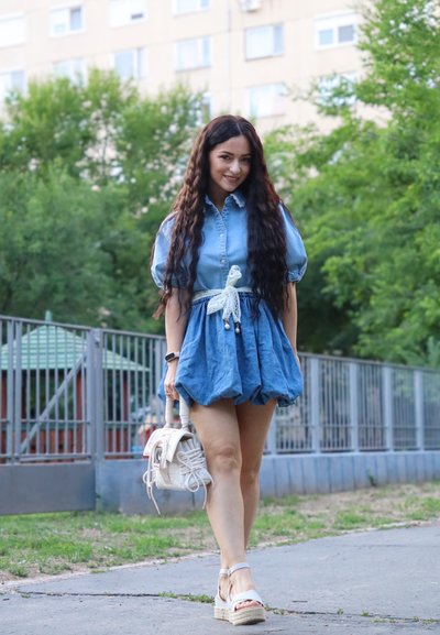 Vestido de mezclilla con mangas abullonadas, falda fruncida y cinturón blanco. Sandalias de cuña; sosteniendo un pequeño bolso blanco. Árboles verdes y parque infantil de fondo.