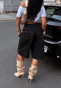 Shorts di jeans neri con cuciture a contrasto, abbinati a stivali beige oversize. Una borsa a tracolla marrone con motivo è indossata sulla spalla.