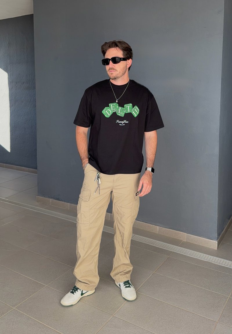 Man draagt een zwart grafisch T-shirt, beige cargobroek, witte sneakers, zwarte zonnebril en ketting, staat binnen tegen een grijze muur.