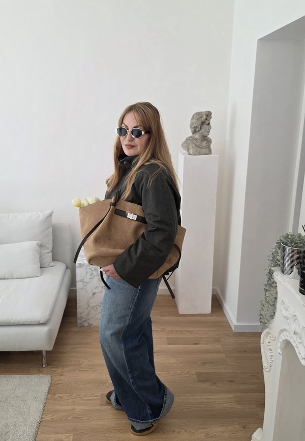 Femme aux longs cheveux et lunettes de soleil se tient à l'intérieur, tenant un sac fourre-tout beige avec des fleurs blanches, près d'un canapé blanc et d'une sculpture de buste classique.