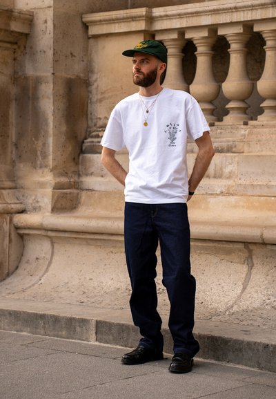 Camiseta blanca de algodón con estampado gráfico negro, combinada con jeans azul oscuro y zapatos negros. Lleva una gorra verde y joyas en capas.