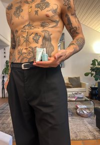 Dwa produkty do pielęgnacji skóry marki ELEMIS: jasnoniebieska tubka i srebrna buteleczka z kroplomierzem, trzymane w dłoni na tle tatuaży i wnętrza pomieszczenia.