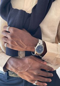 Persoană cu pielea închisă la culoare, purtând o cămașă bej și un pulover bleumarin legat în jurul umerilor, cu un ceas de argint și mai multe inele de argint pe mâinile încrucișate.