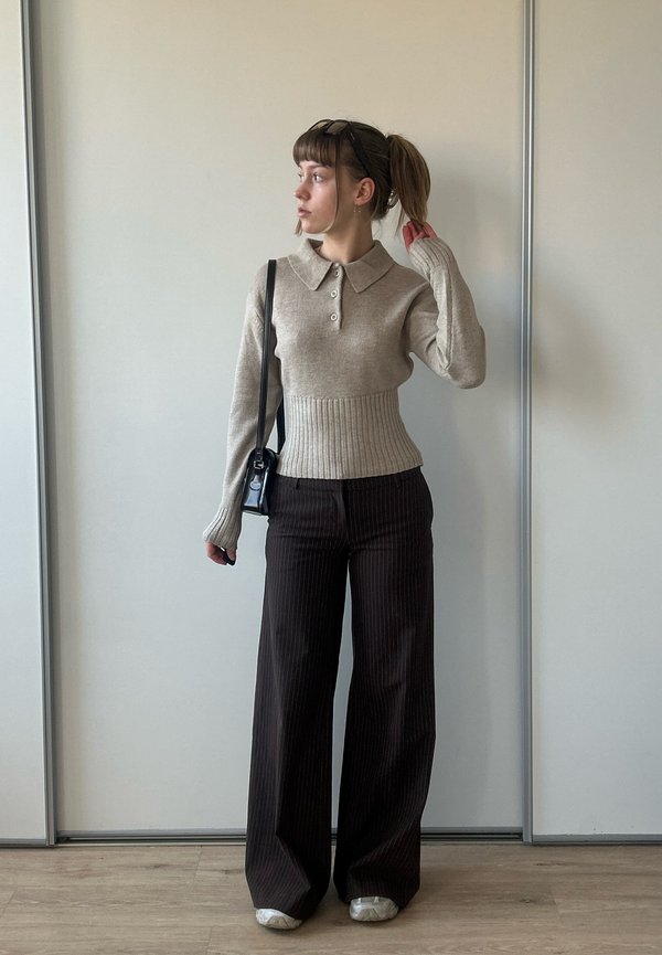 Jonge vrouw met paardenstaart die een beige trui met knopen draagt, brede donkere krijtstreepbroek, witte schoenen en een zwarte schoudertas, staand binnen.