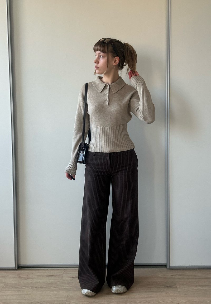 Jeune femme avec queue de cheval portant un pull beige boutonné, un pantalon large à fines rayures foncées, des chaussures blanches et un sac à épaule noir, debout à l'intérieur.