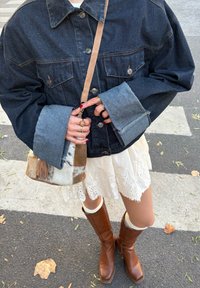 Casaquinho oversized de denim escuro com botões de metal, combinado com uma saia branca com rendas, botas castanhas até ao joelho e uma mala de múltiplos texturas.