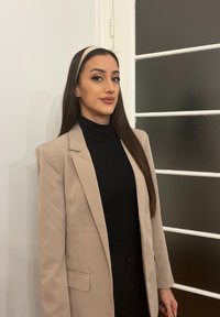 Femme aux longs cheveux foncés portant un blazer beige et un col roulé noir, debout près d’un mur blanc et d’une porte avec des panneaux de verre dépoli.