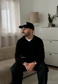 Pull noir et casquette, assis sur un canapé beige clair, pantalon foncé, montre argentée avec des bagues. Chambre moderne avec éclairage doux et décoration.