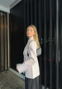 Blazer rose clair à coupe ajustée, sac à main noir et jupe noire, sur fond de barres verticales sombres et d'architecture moderne.