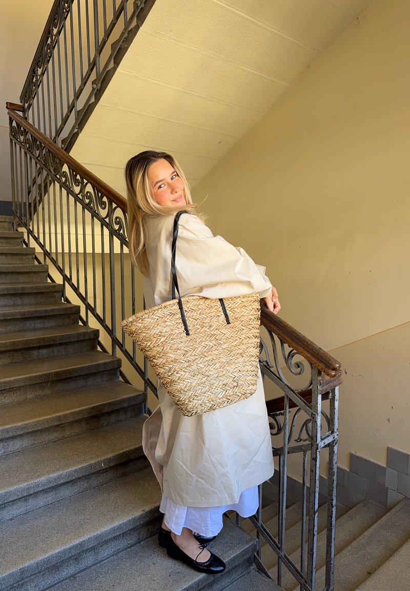 Grande bolsa de palha trançada com alças de couro preto, apresentando um padrão de espinha de peixe, segurada por uma pessoa em uma escada.