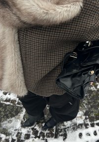 Manteau marron à motif pied-de-poule avec col en fourrure, associé à un sac à main noir brillant et un pantalon noir, dans un décor extérieur enneigé.