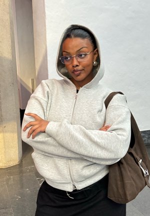 Mujer con gafas y sudadera gris con capucha, con los brazos cruzados, de pie en el interior frente a una pared blanca, llevando un bolso marrón al hombro.