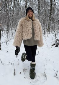Veste courte en fausse fourrure beige, pull kaki, leggings noirs, bottes de neige vertes avec accents blancs, sac à main noir, fond de forêt enneigée.
