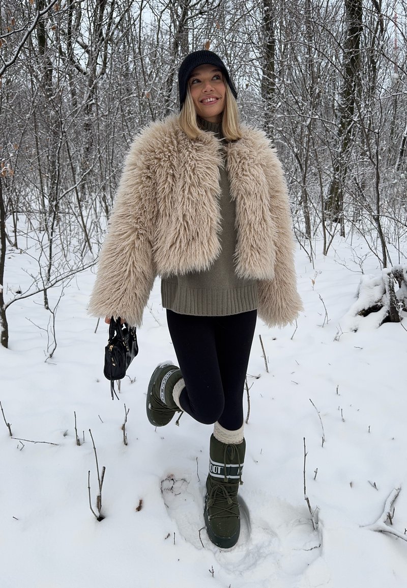 Veste courte en fausse fourrure beige, pull kaki, leggings noirs, bottes de neige vertes avec accents blancs, sac à main noir, fond de forêt enneigée.