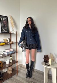 Denim jakna z velikimi žepi, temno modre barve, s prevelikim krojem; v kombinaciji s črnimi kratkimi hlačami z naborki in črnimi čevlji s peto do kolen.
