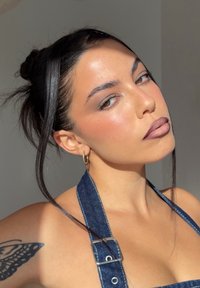 Joven con el cabello oscuro recogido en un moño, lleva tirantes de mezclilla y pendientes de aro dorados, con un tatuaje de mariposa en el hombro, iluminada por luz natural.